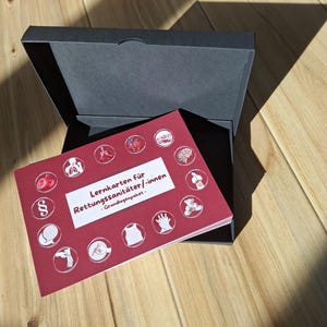 Könnte beinhalten: Ein rotes Karteikartenset mit medizinischen Illustrationen und deutschem Text, betitelt "Lernkarten für Rettungssanitäter/innen - Grundlagenpaket", wird in einer offenen schwarzen Box präsentiert. Die Box steht auf einer Holzoberfläche.