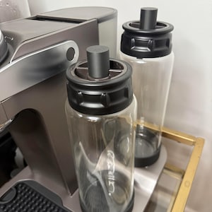 May include: Two clear glass bottles with black lids and black rubber stoppers. The bottles are sitting on a gray surface with a black rubber mat.