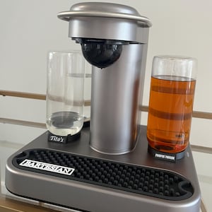May include: A silver countertop cocktail dispenser with a black top and a clear glass container. The dispenser is on a black base with a rubber mat and the words "BARTESIAN" and "PROPER TWELVE" printed on it. Two clear glasses with labels that say "Tito's" and "PROPER TWELVE" are on the base.
