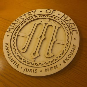 May include: A round, wooden Ministry of Magic emblem. The emblem features the text "Ministry of Magic" and "Ignorantia Juris Non Excusat" in a circular pattern. The center features a stylized "M".