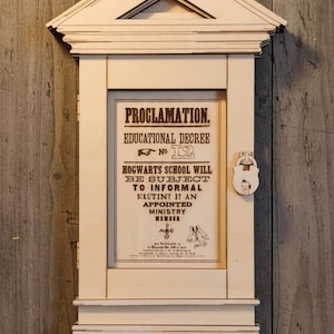 Può includere: Un'applique in legno a forma di casa, con una proclamazione incorniciata. Il testo recita: "PROCLAMAZIONE. DECRETO EDUCATIVO N. 12 LA SCUOLA DI HOGWARTS SARÀ SOGGETTA A CONTROLLI INFORMALI DA UN MEMBRO DEL MINISTERO NOMINATO."