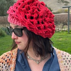May include: A red and pink crocheted beanie with a lacy design. The beanie is worn by a person with dark hair and sunglasses.