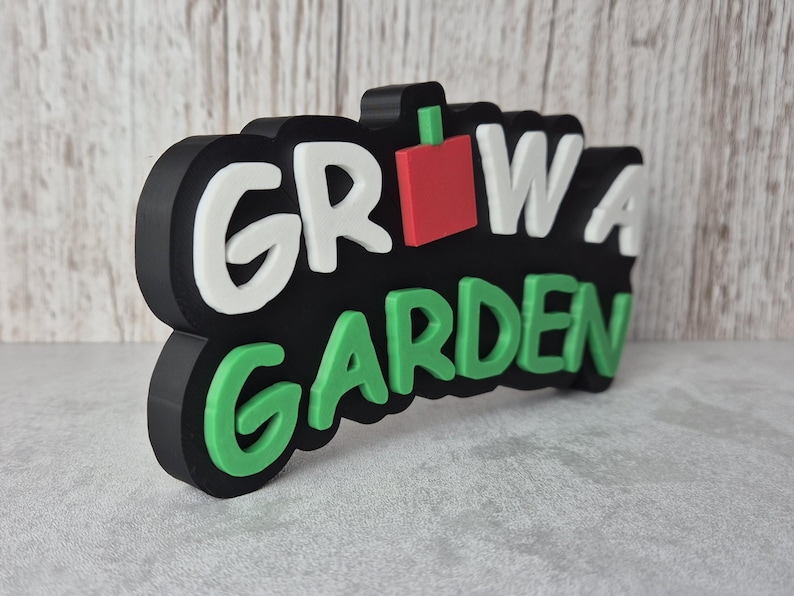 Puede incluir: Un letrero negro con las palabras "GROW A GARDEN" en blanco y verde, con un cuadrado rojo y un acento de tallo verde. El letrero es tridimensional y parece estar hecho de pl&aacute;stico o un material similar. El fondo es una superficie gris claro.