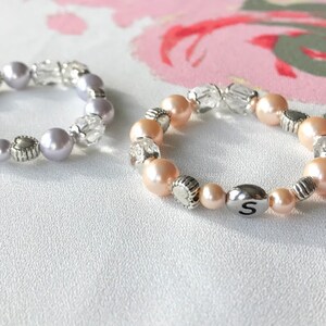 May include: Two beaded bracelets with silver charms and initial letters "G" and "S". The bracelet on the left has light purple beads and clear beads. The bracelet on the right has light pink beads and clear beads.