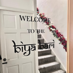 May include: A white door with black lettering that reads "WELCOME TO THE biya bari". A staircase with grey carpet and a floral garland of red and white flowers is visible.