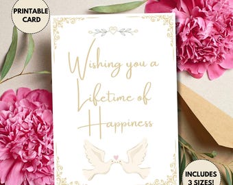 Op de afbeelding: Een witte printbare kaart met de gouden tekst "Wishing you a Lifetime of Happiness". Twee witte duiven met een roze hart zijn afgebeeld. De kaart is omgeven door roze bloemen en groen.