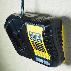 Puede incluir: Cargador de batería de iones de litio DCB1104 12V/20V negro y amarillo. El cargador tiene forma rectangular con una etiqueta amarilla que muestra el voltaje y el estado de carga. El cargador está montado sobre una superficie marrón claro.