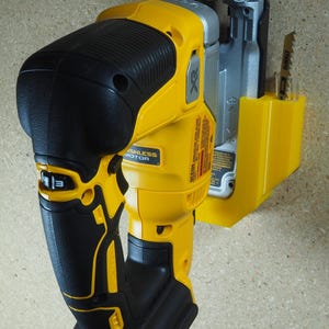 May include: A yellow and black cordless reciprocating saw. The saw features a black handle and a yellow body with the text "DEWALT" and "BRUSHLESS MOTOR". A saw blade is inserted in the front.