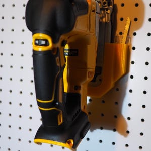 May include: A yellow and black cordless jigsaw with a black handle and a battery pack. The jigsaw is hanging on a white pegboard.
