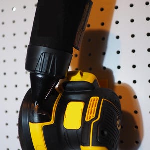 May include: Black and yellow power tool with a cylindrical black attachment. The tool has a rounded base and a black and yellow body. The tool is hanging on a white pegboard.