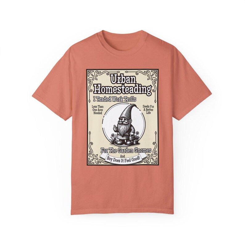 May include: A coral coloured t-shirt featuring a vintage-style illustration of a garden gnome. The illustration is surrounded by a decorative frame and includes the text "Urban Homesteading I Traded Work Trolls Less Than One Acre Needed Seeds For A Better Life For The Garden Gnomes And Boy Does It Feel Good!"