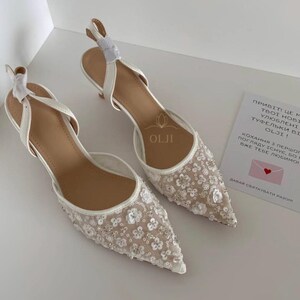 Puede incluir: Un par de zapatos de novia blancos con un delicado bordado floral y una punta puntiaguda. Los zapatos tienen un tacón pequeño y una delicada correa de tobillo.