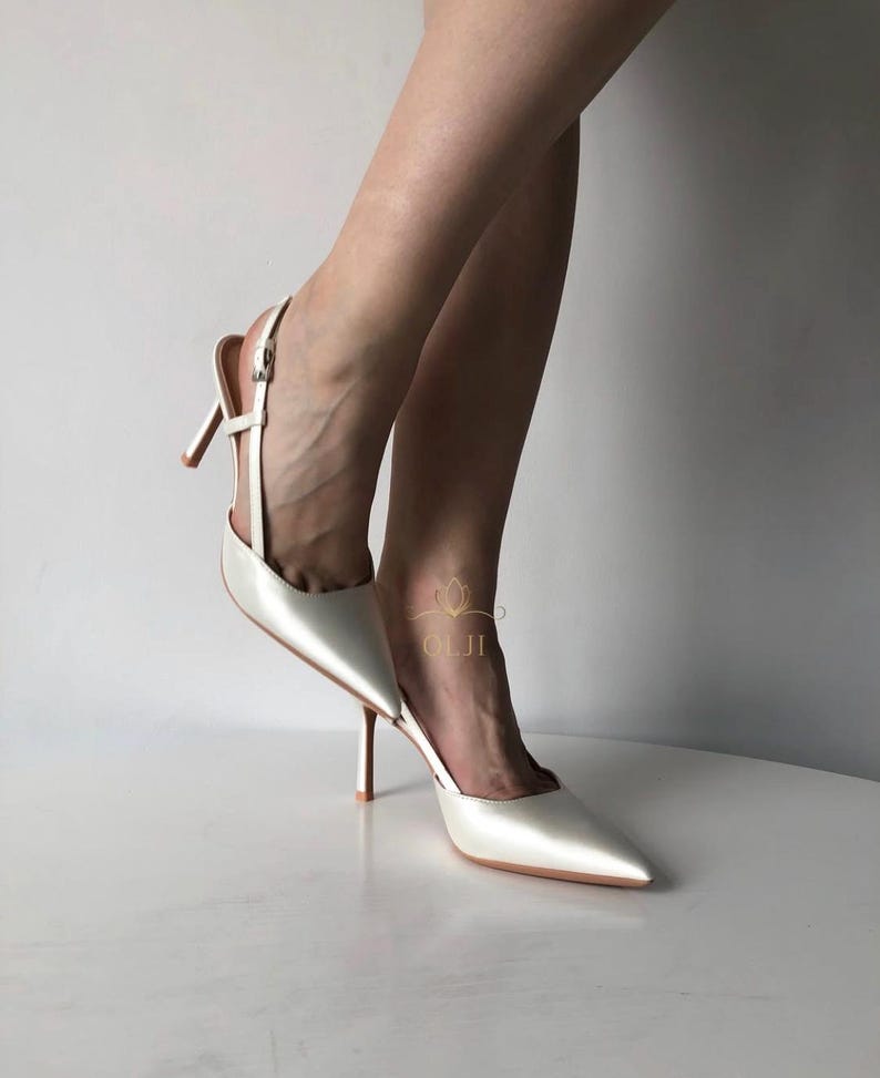 Puede incluir: Zapatos de tac&oacute;n blancos de sat&eacute;n con punta y tacones de aguja. Los zapatos tienen una correa en el tobillo y una altura de tac&oacute;n de aproximadamente 7,6 cm. El dise&ntilde;o es elegante y adecuado para ocasiones formales.