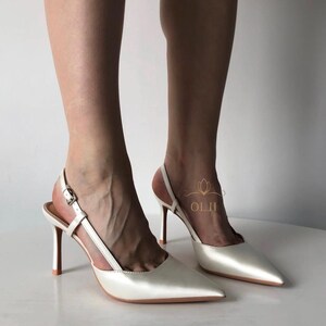 Puede incluir: Zapatos de tac&oacute;n blancos de sat&eacute;n con punta y tacones de aguja. Los tacones tienen un cierre de hebilla y una correa que rodea el tobillo. Los tacones miden aproximadamente 10 cm de alto.
