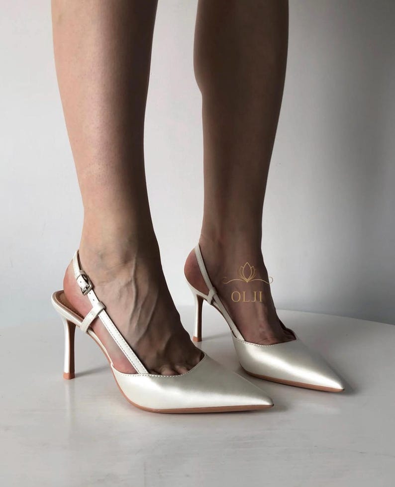 Puede incluir: Zapatos de tac&oacute;n blancos de sat&eacute;n con punta y tacones de aguja. Los tacones tienen un cierre de hebilla y una correa que rodea el tobillo. Los tacones miden aproximadamente 10 cm de alto.