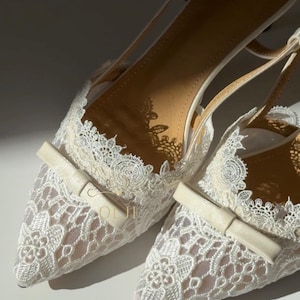May include: A pair of elegant white lace slingback heels. The shoes feature a pointed toe, delicate lace detailing, and a small bow accent. The heels have a slender strap and a low heel, perfect for special occasions.