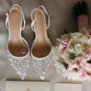 Puede incluir: Un par de zapatos de novia blancos con adornos de cristal, un ramo de flores rosas y blancas, dos anillos de boda dorados y una invitación con texto. Los zapatos tienen punta y correas en el tobillo.