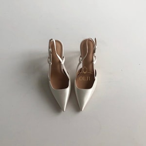 Puede incluir: Un par de zapatos de tac&oacute;n slingback de color marfil con punta. Los zapatos tienen correas finas y hebillas para ajustar. El nombre de la marca "OLJI" es visible dentro de uno de los zapatos. Los tacones est&aacute;n sobre un fondo blanco.