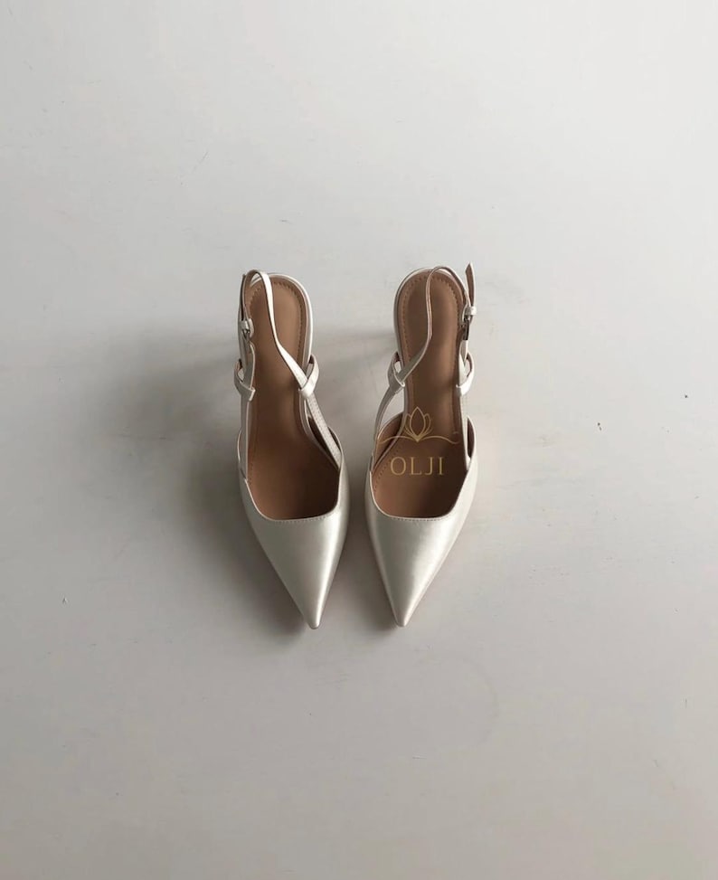 Puede incluir: Un par de zapatos de tac&oacute;n slingback de color marfil con punta. Los zapatos tienen correas finas y hebillas para ajustar. El nombre de la marca "OLJI" es visible dentro de uno de los zapatos. Los tacones est&aacute;n sobre un fondo blanco.