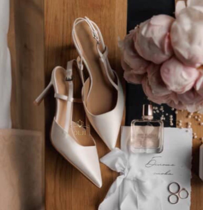 Puede incluir: Zapatos de tac&oacute;n slingback de sat&eacute;n marfil con puntera y tacones de aguja. Los zapatos se exhiben con un frasco de perfume, un ramo de flores rosas y anillos de boda sobre una superficie de madera.