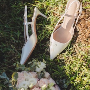 Puede incluir: Un par de zapatos de tac&oacute;n blancos con punta y tacones de aguja, sobre hierba verde. Un ramo de rosas blancas y rosas y vegetaci&oacute;n est&aacute; en primer plano. Los tacones tienen un delicado dise&ntilde;o de correas.