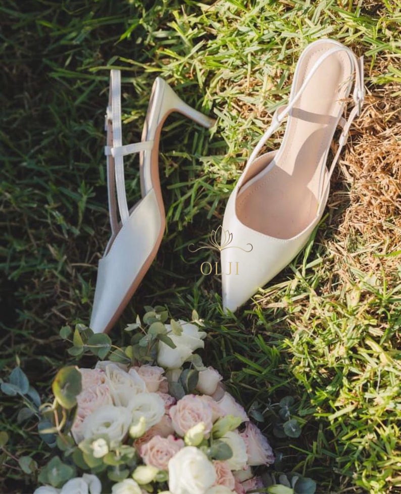 Puede incluir: Un par de zapatos de tac&oacute;n blancos con punta y tacones de aguja, sobre hierba verde. Un ramo de rosas blancas y rosas y vegetaci&oacute;n est&aacute; en primer plano. Los tacones tienen un delicado dise&ntilde;o de correas.
