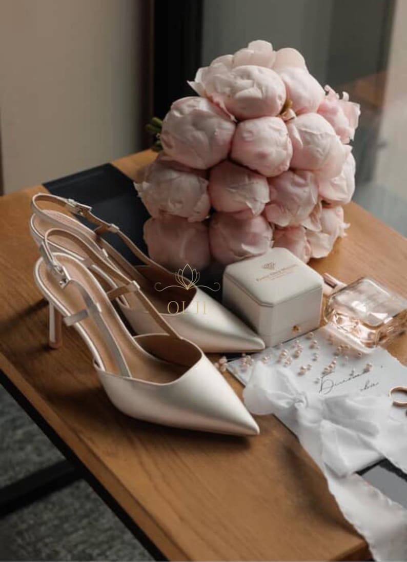 Puede incluir: Un par de zapatos de tac&oacute;n slingback de sat&eacute;n marfil con punta, junto a un ramo de peon&iacute;as rosas. Una peque&ntilde;a caja blanca, perfume y una nota manuscrita tambi&eacute;n est&aacute;n sobre la superficie de madera.