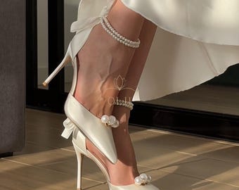 Op de afbeelding: Witte satijnen hoge hakken met parelversiering en een strikdetail. De schoenen hebben een spitse neus en een dunne stilettohak.