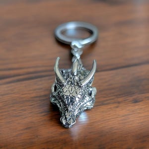 May include: Silver dragon head keychain with a detailed textured design. The dragon has a long snout and two horns.
