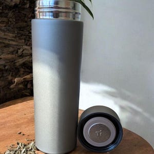 May include: A gray insulated tumbler with a silver lid and a black cap. The cap is unscrewed and placed next to the tumbler. The tumbler is on a wooden surface with dried herbs scattered around it.