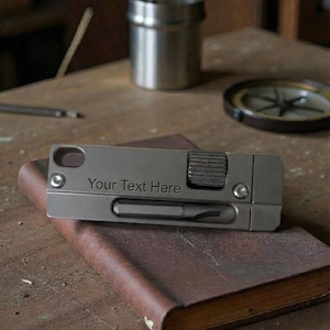 Può includere: Un multiutensile grigio con la scritta "Your Text Here" incisa, appoggiato su un libro rilegato in pelle marrone. Un piccolo contenitore di metallo e una bussola sono sullo sfondo, il tutto su una superficie di legno.