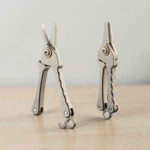 May include: Two silver-toned metal multi-tool pliers with a matte finish. One tool is shown in an open position with pointed jaws, while the other is closed. Both feature a central brass-colored pivot screw and textured handles with a loop at the base.