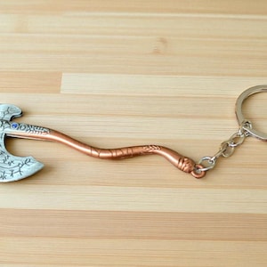 May include: A metal axe keychain with a silver axe head and a copper-colored handle. The axe head features an engraved design and a small blue gem. The keychain includes a silver ring and chain.