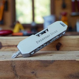 May include: A silver multi-tool with the text "Everyday Duke" engraved on the side. The tool has a screwdriver bit extended and is resting on a wooden surface. The background is a blurred workshop setting.