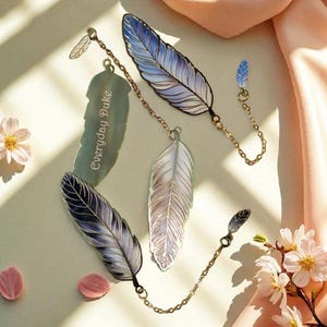 May include: Feather-shaped bookmarks in shades of blue, gray, and white, connected by delicate gold chains. One bookmark has the text "Everyday Duke". The image is styled with flowers and a peach-colored fabric.