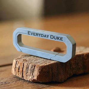 Pode incluir: Um porta-chaves retangular cinza com o texto "EVERYDAY DUKE" gravado. O porta-chaves está sobre um pedaço de madeira castanha áspera, com um fundo desfocado.