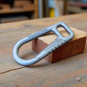 May include: A silver-colored bottle opener with the text "EVERYDAY DUKE" is displayed on a wooden surface. The opener has a curved design with a rectangular opening and a series of diagonal lines. It rests on a small, rectangular piece of wood.