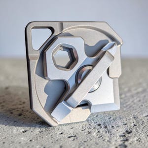May include: A silver multi-tool with various integrated tools, including a hex wrench, screwdriver, and other functional components. The tool has a compact, square design with a key ring hole. The tool is made of metal and sits on a concrete surface.
