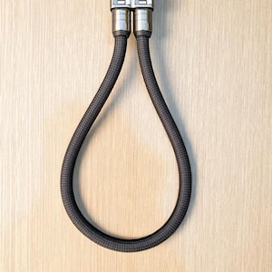 Op de afbeelding: Een zwart en zilveren sleutelhanger met een lusontwerp. De sleutelhanger heeft een metalen sluiting en een geweven zwart koord dat een druppelvorm vormt. De achtergrond is een lichte houtnerf.