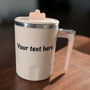 May include: A cream-colored travel mug with a lid and handle. The mug has a silver band around the top and a light pink lid with a small tab. The text "Your text here" is printed on the front of the mug.