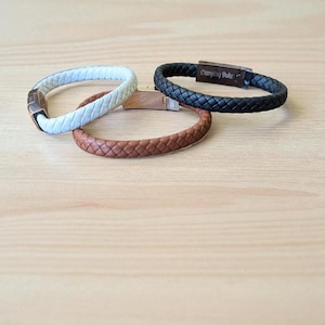 Könnte beinhalten: Drei Lederarmbänder in Weiß, Braun und Schwarz, jeweils mit geflochtenem Design. Das schwarze Armband trägt den Text "Everyday Duke". Die Armbänder werden auf einer hellen Holzoberfläche präsentiert.