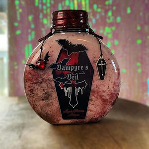 May include: A round, clear bottle with a dark red cap and a label that reads "Vampyre's Veil." The bottle is decorated with bat and coffin charms. The liquid inside is pink with red flecks, creating a spooky aesthetic.