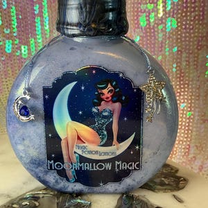 Könnte beinhalten: Eine runde, hellblaue Flasche mit dunklem Verschluss und einem dekorativen Etikett mit einer Frau auf einem Mond. Das Etikett trägt die Aufschrift "Magic Potion Lotions" und "Moonmallow Magic". Silberne Anhänger hängen an der Flasche.