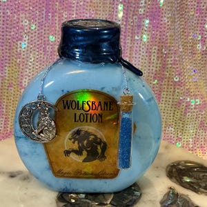 May include: A light blue, round bottle labeled "WOLFSBANE LOTION" with a werewolf illustration. The bottle has a dark blue cap and a silver charm with a wolf and crescent moon. A small glass vial filled with blue glitter hangs from the bottle.