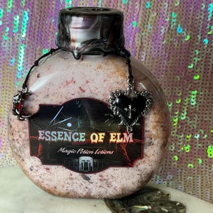 May include: A round, textured bottle with a dark, ornate stopper and a black label that reads "ESSENCE OF ELM Magic Potion Lotions." The bottle is adorned with a black heart pendant and a crescent moon charm.