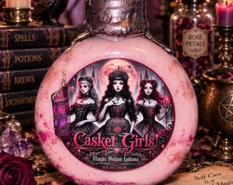 May include: A round, light pink lotion bottle with a metallic cap and a purple crystal charm. The label features three figures in dark dresses, the text "Casket Girls" and "Magic Potion Lotions," and a volume of 10 fl oz (296 ml) printed on the front.