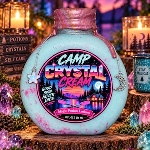 May include: A light blue, round bottle labeled Camp Crystal Cream with a pink marbled cap. The label features a sunset lake scene, the text Good Skin Never Dies, and Magic Potion Lotions. A silver chain with a star and moon charm hangs from the neck.