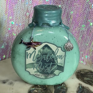 May include: A round, pale blue bottle with a teal cap and a label reading "Megalodon Mist". The bottle is adorned with a shark charm and a silver charm. The background is a shimmering, iridescent fabric.