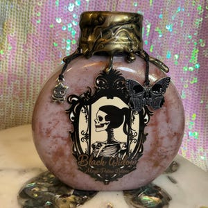 May include: A decorative, pink potion bottle with a bronze-colored top and a black and white label featuring a skeleton. The bottle has a skull charm and a butterfly charm. The text on the label reads "Black Widow Magic Potion Lotions."