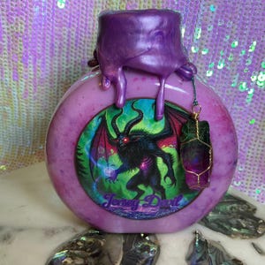 May include: A round, pink bottle with a purple top and dripping accents. The bottle features a circular image of a horned demon with the text "Jersey Devil". A wire-wrapped crystal hangs from the bottle.